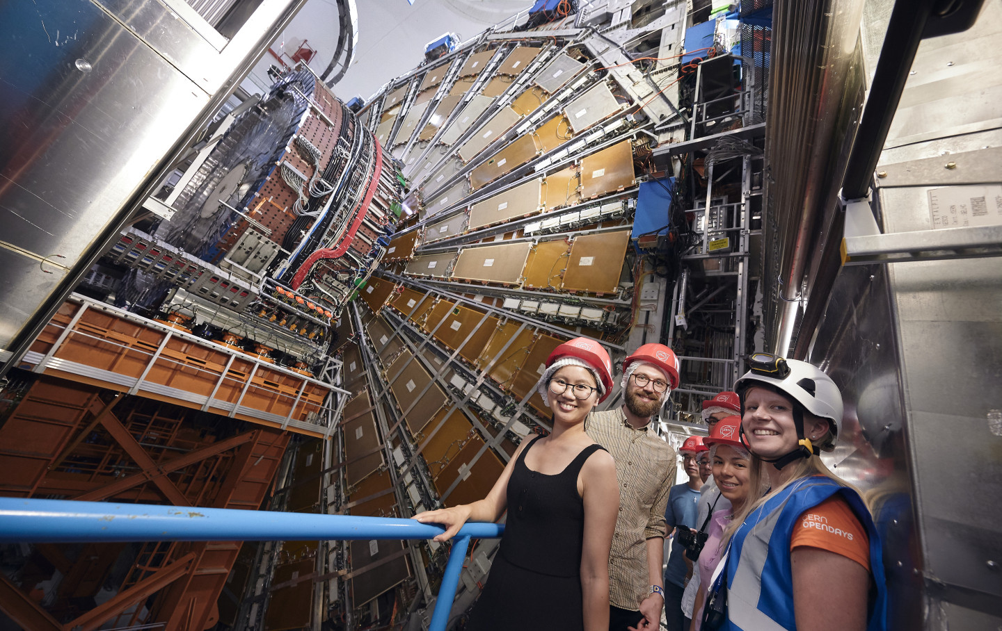 A group of men and women in hardhats pose, smiling, by a blue railing separating them from a large, complex particle detector at a high-energy accelerator laboratory
